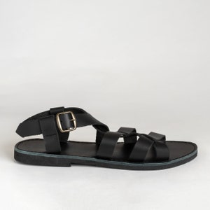 May include: A black flat sandal featuring multiple wide straps across the foot and an adjustable ankle strap with a metallic gold-toned buckle. The footwear has a flat sole and a simple design with a secure closure system for the ankle area.