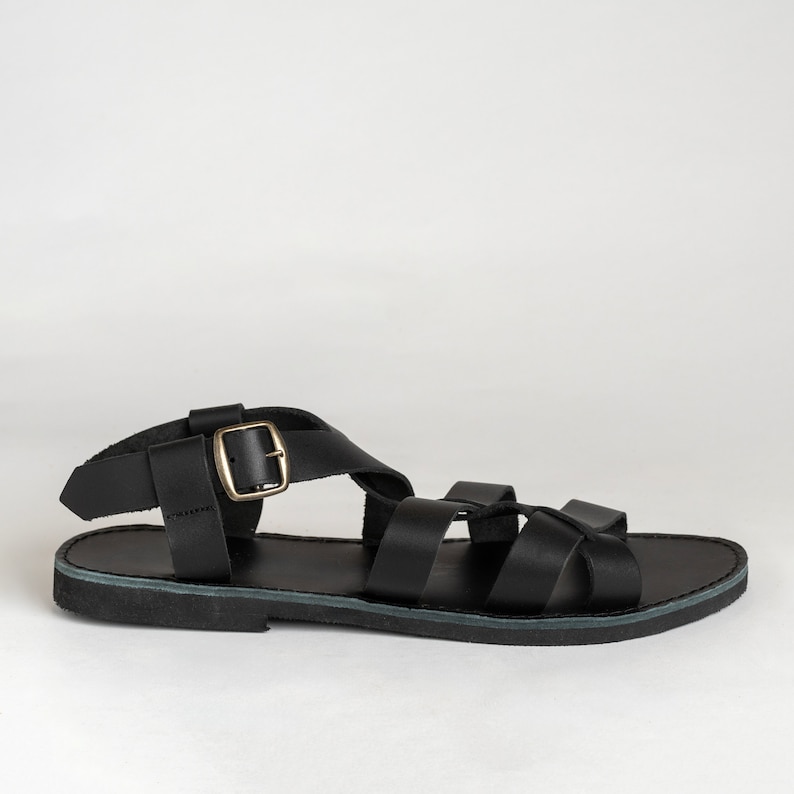 May include: A black flat sandal featuring multiple wide straps across the foot and an adjustable ankle strap with a metallic gold-toned buckle. The footwear has a flat sole and a simple design with a secure closure system for the ankle area.