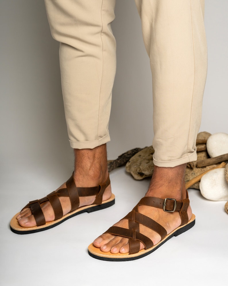 May include: A pair of dark brown leather sandals featuring multiple crisscrossing straps across the top of the foot. The sandals include a side buckle closure and a flat black sole. The design consists of wide leather bands that secure the foot.