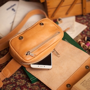 Peut inclure: Une banane en cuir de couleur beige avec une poche zipp&eacute;e et un t&eacute;l&eacute;phone &agrave; l'int&eacute;rieur. La banane est pos&eacute;e sur une surface brune et verte.
