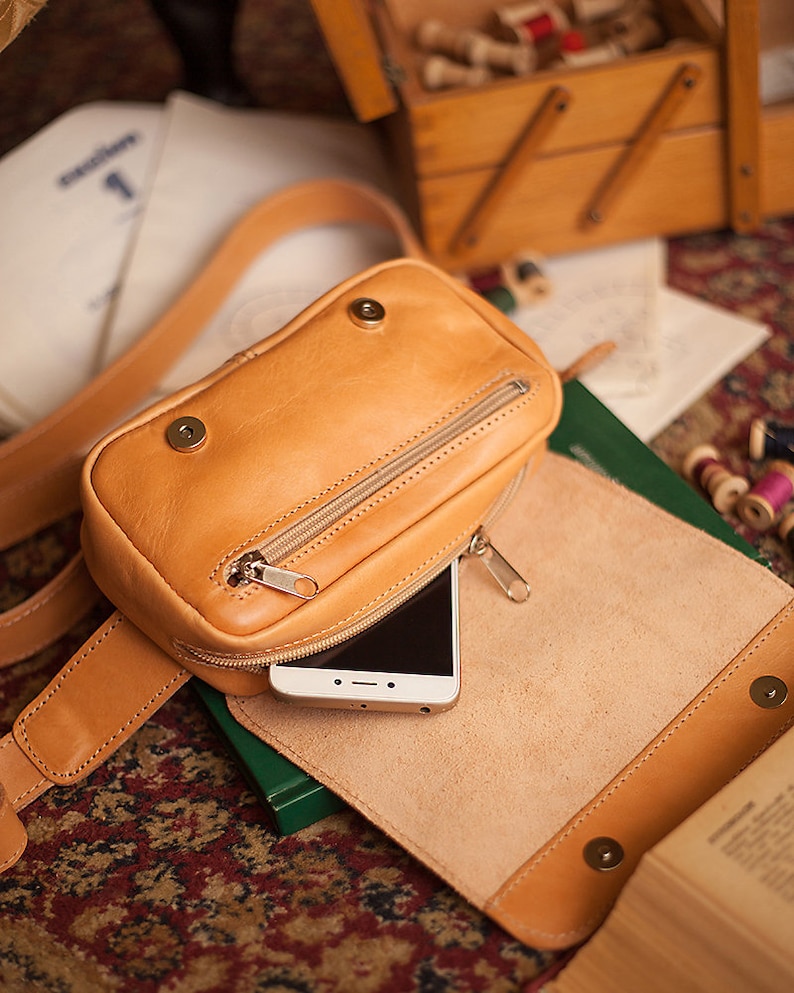 Peut inclure: Une banane en cuir de couleur beige avec une poche zipp&eacute;e et un t&eacute;l&eacute;phone &agrave; l'int&eacute;rieur. La banane est pos&eacute;e sur une surface brune et verte.