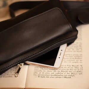 May include: Black leather fanny pack with a zippered compartment, partially open to reveal a white smartphone. The bag rests on an open book with Greek text. The zipper is gold-colored.
