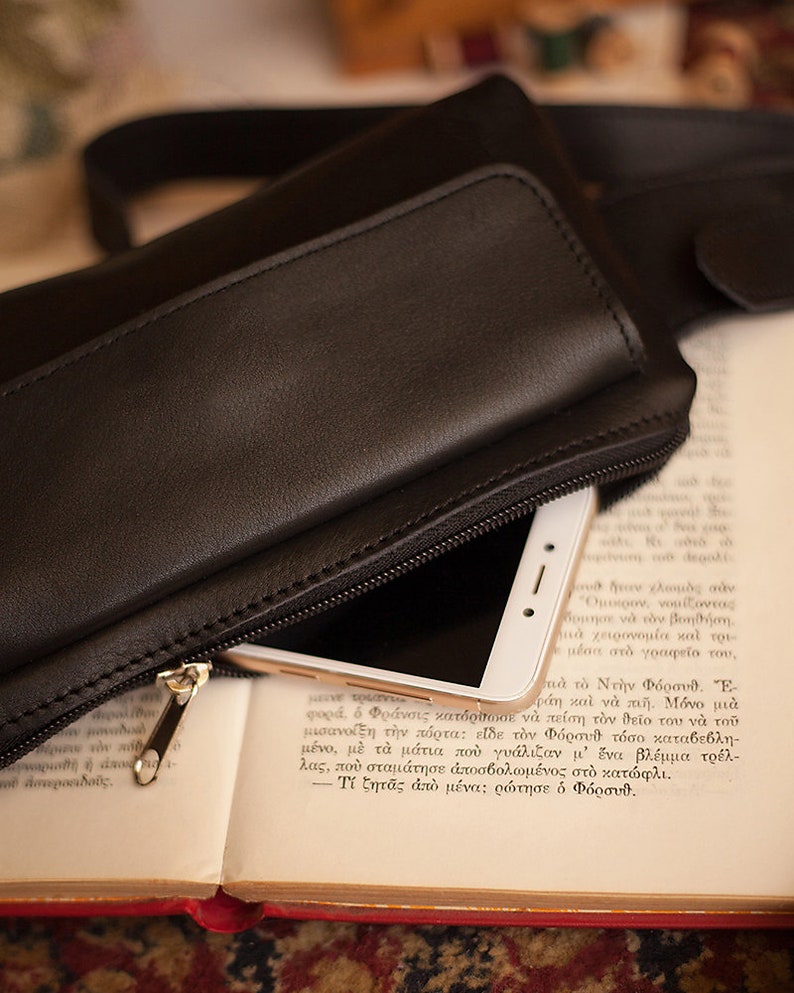 May include: Black leather fanny pack with a zippered compartment, partially open to reveal a white smartphone. The bag rests on an open book with Greek text. The zipper is gold-colored.