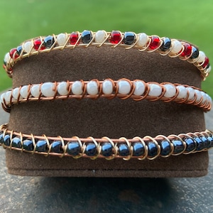 May include: Three wire-wrapped bracelets with different colored beads. The top bracelet has red, white, and black beads. The middle bracelet has white beads. The bottom bracelet has black beads.