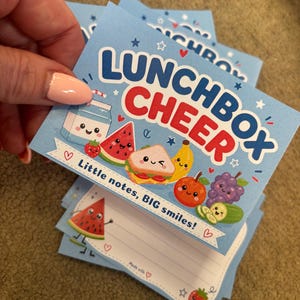 May include: Light blue Lunchbox Cheer notepads featuring illustrations of smiling food items such as watermelon, sandwiches, and bananas. The notepad has the text "Lunchbox Cheer" and "Little notes, BIG smiles!" printed on it.