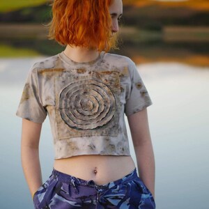 Puede incluir: Una camiseta gris claro recortada con un diseño circular en tonos marrones, verdes y blancos. El diseño se asemeja a un tronco de árbol con anillos. La camiseta la lleva puesta una persona con el pelo rojo.