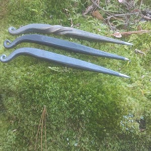May include: Three hand-forged metal tools with a dark finish rest on a bed of green moss. Each tool has a curved handle with a spiral or looped end and a pointed tip. The tools appear to be made of iron or steel.