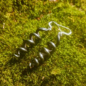 May include: A pair of dark gray spiral earrings with silver-colored hooks, resting on a bed of green moss. The earrings have a twisted, coiled design, adding a unique touch to the jewelry.