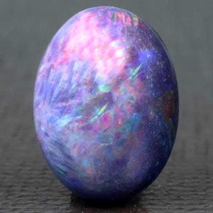 May include: A polished, oval-shaped gemstone with a swirling pattern of colors. The stone features a blend of purple, pink, and blue hues, with iridescent flashes of green and yellow. The overall effect is a galaxy-like appearance, suggesting an opal.