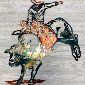 May include: Metal wall art sculpture of a cowboy riding a bucking bull. The sculpture is made of metal and has a rustic, weathered look. The cowboy is wearing a hat and boots, and the bull is bucking its head and legs.