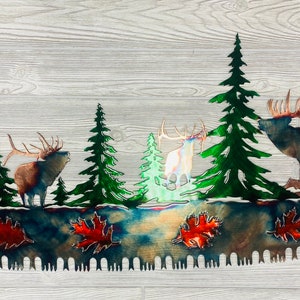 May include: Metal wall art depicting a rustic cabin, pine trees, and a family of elk. The art is shaped like a saw blade with a distressed metal finish.