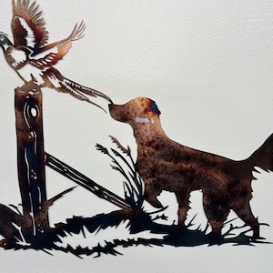 Może przedstawiać: Metalowa ozdoba ścienna przedstawiająca brązowego psa z przepiórką w locie. Pies stoi na trawiastym terenie z tyłu z drewnianym słupkiem. Przepiórka leci nad słupkiem.