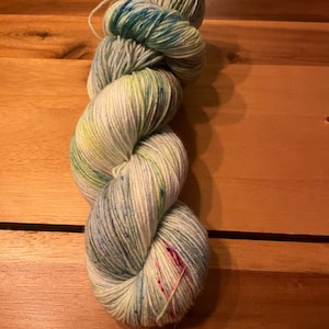 Könnte beinhalten: Eine Garnrolle in einer hellblauen, grünen und weißen Farbkombination mit rosa Sprenkeln. Das Garn ist handgefärbt und hat ein buntes Farbmuster.