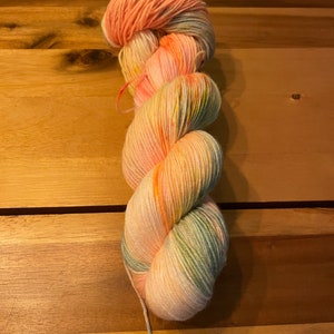 Könnte beinhalten: Eine handgefärbte Garnrolle in den Farben Pfirsich, Rosa, Gelb und Grün. Das Garn ist spiralförmig gedreht.
