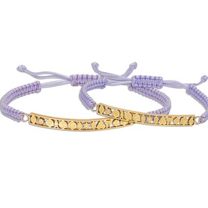 May include: Two purple braided bracelets with a gold charm bar featuring a variety of symbols. The charms are connected to the bracelets with a gold loop.