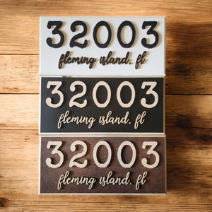 May include: Three address signs stacked on a wooden surface. Each sign displays the number "32003" above the text "fleming island. fl". The top sign is white with black numbers, the middle is black with beige numbers, and the bottom is brown with beige numbers.