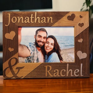 May include: Wooden photo frame with the names "Jonathan & Rachel" engraved. The frame has a two-tone design with heart accents and holds a photo of a smiling couple. The frame is made of wood.