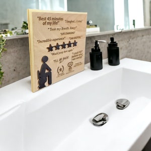 May include: A wooden plaque with a humorous review of a bathroom experience. The plaque features a silhouette of a person sitting on a toilet, five stars, and the text "Best 45 minutes of my life!"  "Took my breath away" "Life changing" "Incredible experience" "Would poop here again" "I'll be back"  "I could not believe what I was seeing!"