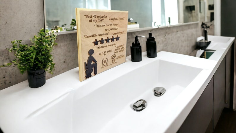 May include: A wooden plaque with a humorous review of a bathroom experience. The plaque features a silhouette of a person sitting on a toilet, five stars, and the text "Best 45 minutes of my life!"  "Took my breath away" "Life changing" "Incredible experience" "Would poop here again" "I'll be back"  "I could not believe what I was seeing!"