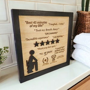 May include: A wooden sign with a black frame features a humorous review of a bathroom experience. The sign includes a cartoon illustration of a person sitting on a toilet, along with text that reads "Best 45 minutes of my life!" and "I laughed... I cried." The sign also includes a five-star rating and the text "Would poop here again."