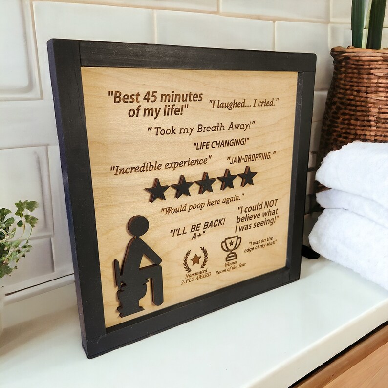 May include: A wooden sign with a black frame features a humorous review of a bathroom experience. The sign includes a cartoon illustration of a person sitting on a toilet, along with text that reads "Best 45 minutes of my life!" and "I laughed... I cried." The sign also includes a five-star rating and the text "Would poop here again."