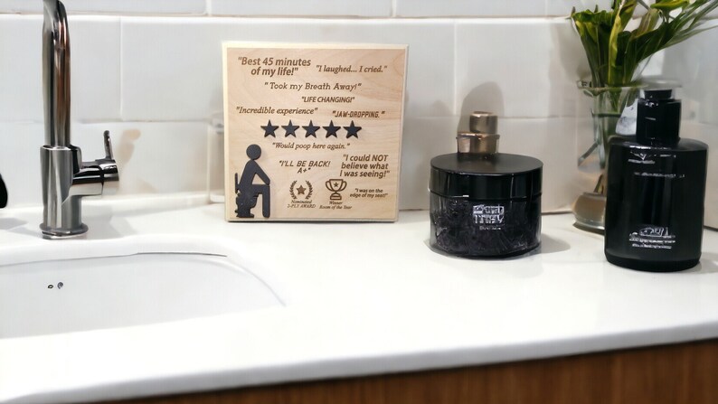 May include: A wooden sign with a humorous review of a bathroom experience. The sign reads "Best 45 minutes of my life!" and includes a five-star rating. The sign is sitting on a white countertop next to a black jar and a black bottle.