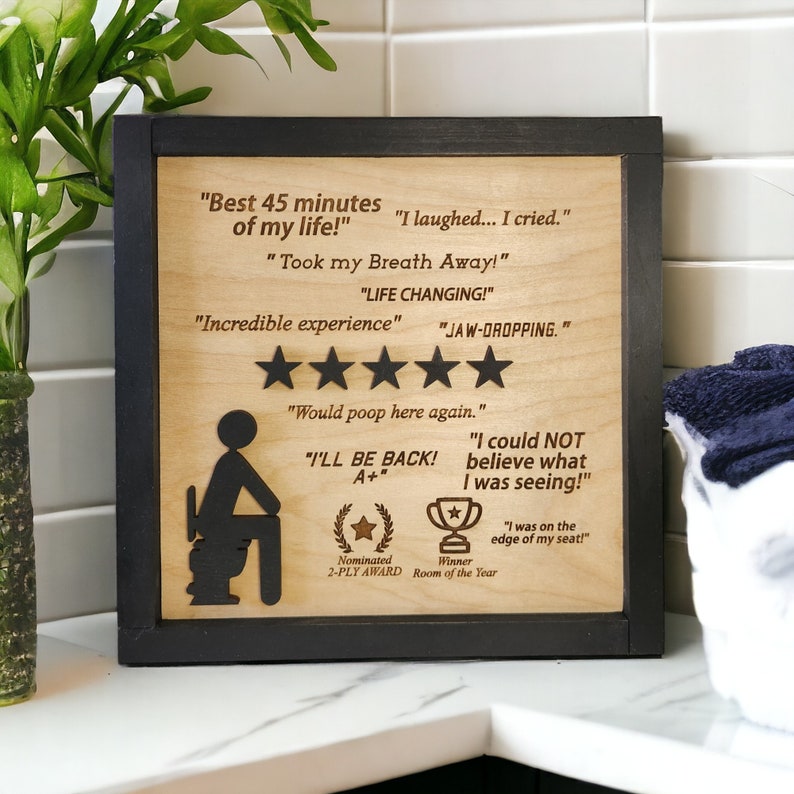 May include: A wooden sign with a black frame features a cartoon illustration of a person sitting on a toilet. The sign includes text that reads "Best 45 minutes of my life!" and "I laughed... I cried." along with a five-star rating and other humorous reviews.