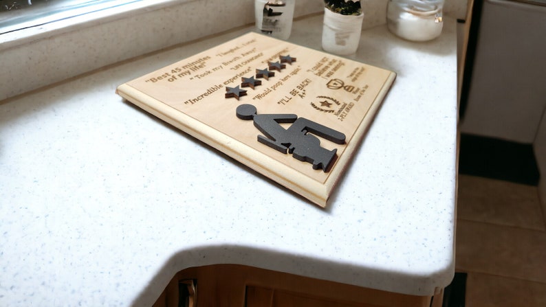 May include: A wooden plaque with a humorous message about a bathroom experience. The plaque features a silhouette of a person sitting on a toilet, five stars, and the text "Best 45 minutes of my life."  "Took my breath away." "Incredible experience." "Would you recommend?" "I'll be back!"  "A+."