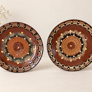 May include: Two round ceramic plates featuring a glossy brown glaze with intricate, hand-painted patterns in cream, black, and dark brown. The designs consist of concentric circles, radiating feather-like motifs, and small dots along the outer rim.