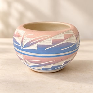 May include: A small, rounded ceramic bowl featuring a pastel colour palette of soft pink and light blue. The exterior surface is decorated with carved, geometric, white-coloured patterns that wrap around the circumference of the vessel.