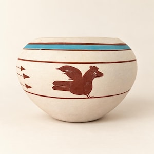 Può includere: Una ciotola in ceramica arrotondata con finitura bianco sporco opaca, caratterizzata da un motivo a uccello dipinto a mano in rosso scuro, strisce orizzontali rosso scuro e una banda orizzontale blu turchese vicino al bordo, con tre piccole frecce sul lato.