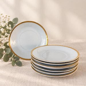 Puede incluir: Un juego de platillos de cerámica blanca con un borde dorado fino y reflectante. Un platillo está colocado en posición vertical mientras que los demás están apilados. El diseño es sencillo y circular, con un acabado liso y borde dorado.