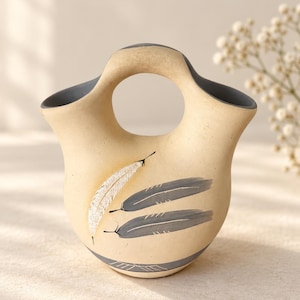 Pode incluir: Um vaso de cerâmica bege com design de bico duplo e alça central. O exterior apresenta uma pena branca pintada e duas penas cinzentas, com uma faixa cinzenta e detalhes de linhas em torno da base e das bordas internas.