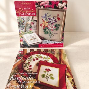 May include: Two Cross Stitch & Needlework Keepsake Calendars from 1999 and 2000. The 1999 calendar cover features a framed floral cross stitch and a journal. The 2000 calendar cover displays a red pillow with a floral cross stitch design.