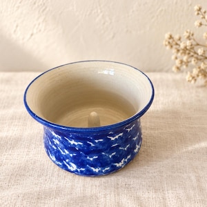 May include: A ceramic butter bell with a cream-colored interior and a small central post. The exterior features a vibrant blue glaze with a white, textured, cloud-like pattern. The rim is finished with a thin, solid blue line around the edge.