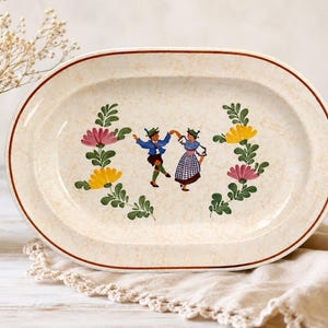May include: An oval ceramic serving platter with a cream-colored speckled finish and a thin brown rim. The center features a painted illustration of two figures dancing, flanked by two symmetrical floral sprays with pink and yellow flowers.