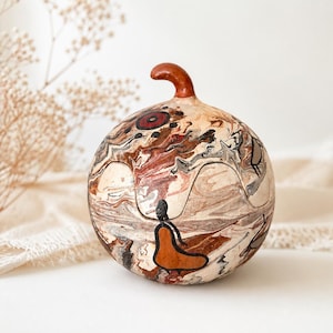 Puede incluir: Un recipiente de calabaza esférico con una superficie lisa y pulida que presenta un patrón jaspeado en colores crema, marrón, negro y óxido. La parte superior tiene un pequeño tallo curvo y el cuerpo está decorado con figuras de líneas negras.