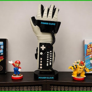 May include: A Power Glove controller with the text "POWER GLOVE" and a black control panel. The image also features Super Mario Bros. video game boxes and figurines of Mario and Bowser. The text "WARP ZONE" is also visible.