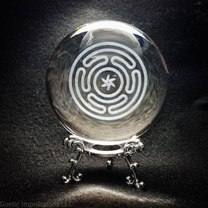 May include: A clear glass orb with a white etched design of a five-pointed star inside a circle surrounded by four concentric circles. The orb is on a silver stand with three legs.