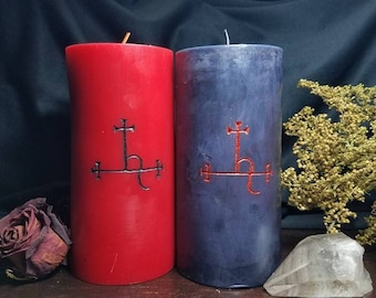 May include: Two cylindrical pillar candles, one bright red and one dark blue, each featuring a black or red engraved sigil symbol on the front. The red candle has a single wick, and the blue candle has a single wick, both standing upright.