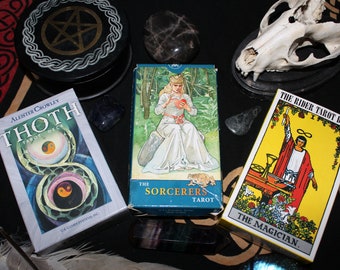 May include: Three tarot card decks, including the Rider Waite Tarot Deck, the Thoth Tarot Deck by Aleister Crowley, and the Sorcerers Tarot Deck. The Rider Waite deck shows the Magician card, the Thoth deck shows the card with a snake and yin yang symbol, and the Sorcerers deck shows a woman in a white dress holding a heart.