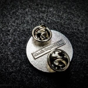 Puede incluir: La parte posterior de un pin circular plateado con un rect&aacute;ngulo negro que dice "Goetic Impressions" en letras blancas. El pin tiene dos cierres met&aacute;licos en la parte posterior.