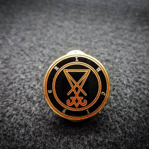 Puede incluir: Un pin de color dorado con fondo negro y un s&iacute;mbolo naranja. El s&iacute;mbolo es un pentagrama con una cruz en su interior. Las letras "LVCIFER" est&aacute;n escritas alrededor del s&iacute;mbolo.