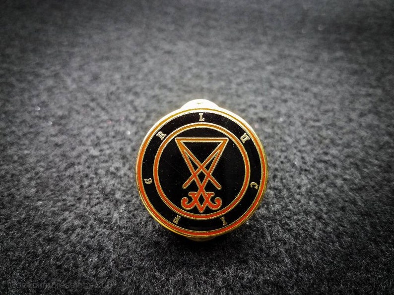 Puede incluir: Un pin de color dorado con fondo negro y un s&iacute;mbolo naranja. El s&iacute;mbolo es un pentagrama con una cruz en su interior. Las letras "LVCIFER" est&aacute;n escritas alrededor del s&iacute;mbolo.
