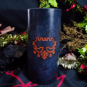 May include: A dark blue pillar candle featuring an orange sigil. The candle is cylindrical in shape. The background includes festive greenery and a dark surface.
