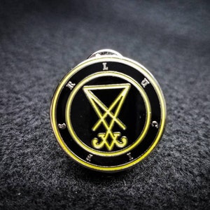 Puede incluir: Un pin de esmalte negro y amarillo con un sigilo dentro de un c&iacute;rculo. El sigilo es un pentagrama con una cruz en su interior. El c&iacute;rculo tiene las letras L, W, I, C, R y S alrededor del exterior.