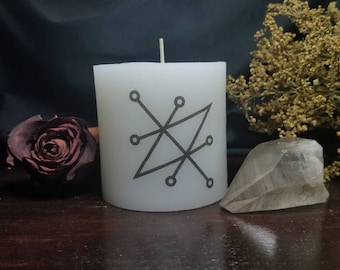 Può includere: Candela bianca con un sigillo nero sul lato. Il sigillo è un disegno geometrico con un triangolo e linee. La candela è appoggiata su una superficie di legno con un cristallo di quarzo trasparente e fiori secchi.