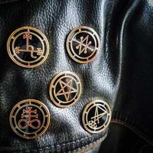 Puede incluir: Cinco pines de metal de color dorado con esmalte negro y detalles rojos. Cada pin presenta un s&iacute;mbolo diferente, incluyendo un pentagrama, una cruz y un c&iacute;rculo con una l&iacute;nea que lo atraviesa.