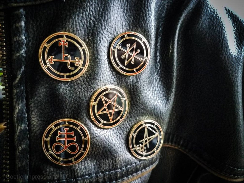 Puede incluir: Cinco pines de metal de color dorado con esmalte negro y detalles rojos. Cada pin presenta un s&iacute;mbolo diferente, incluyendo un pentagrama, una cruz y un c&iacute;rculo con una l&iacute;nea que lo atraviesa.