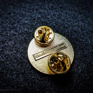 Puede incluir: Pin de metal de color dorado con un dise&ntilde;o redondo. La parte posterior del pin tiene el texto "Goetic Impressions" impreso.
